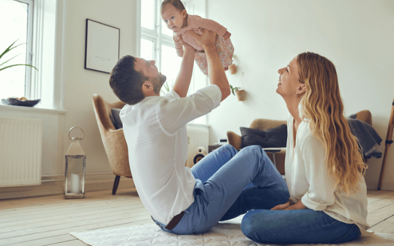 A photo of a father holding a baby in the air beside a mother. Both are sitting on the floor of a living room.