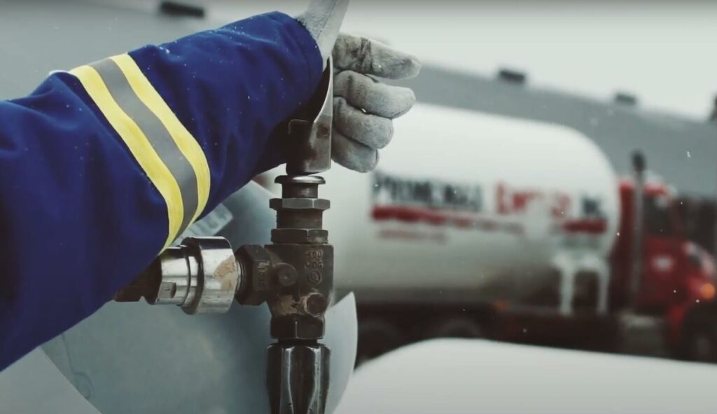 A photo of a hand with gloves filling a propane tank, with a Primemax Energy fuel truck in the background.