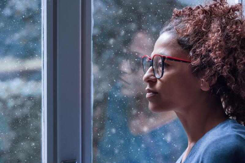 A photo of a woman wearing glasses looking out the window of her home in the winter. Snow is on the window.